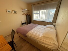 Una camera da letto con un letto, una sedia, un termosifone, una finestra con tende e un appendiabiti con grucce.