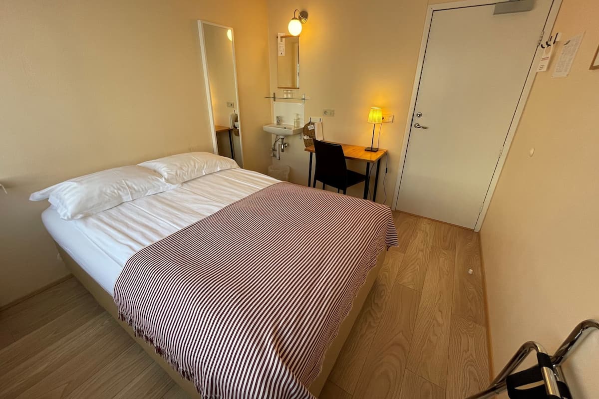 Quarto de hotel com cama, escrivaninha, cadeira, espelho, pia e porta.