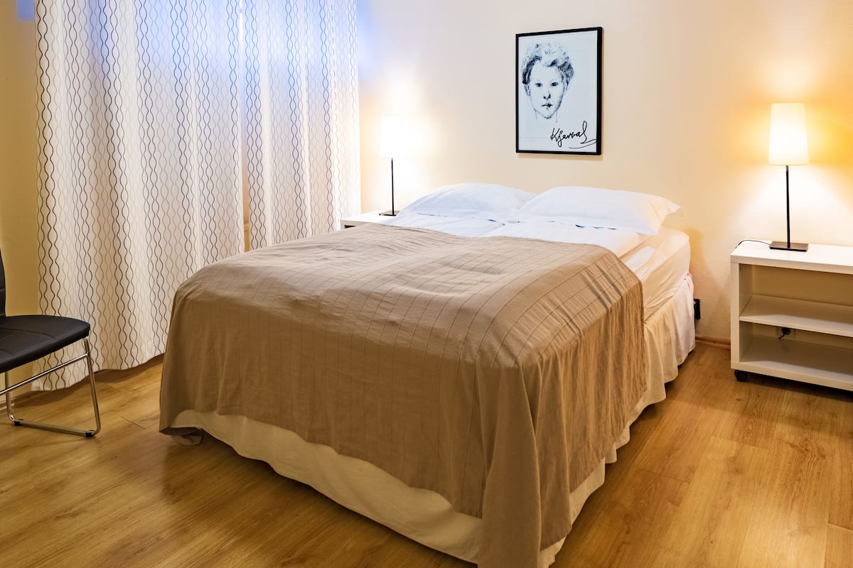 Uma cama bem arrumada com um cobre-leito bege, uma mesa de cabeceira com abajur e um quadro pendurado na parede.
