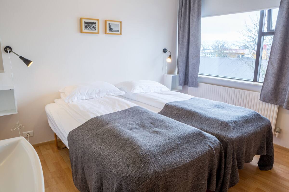 Quarto de hotel com uma cama, duas mesas de cabeceira, uma janela com cortinas e um abajur fixado na parede.