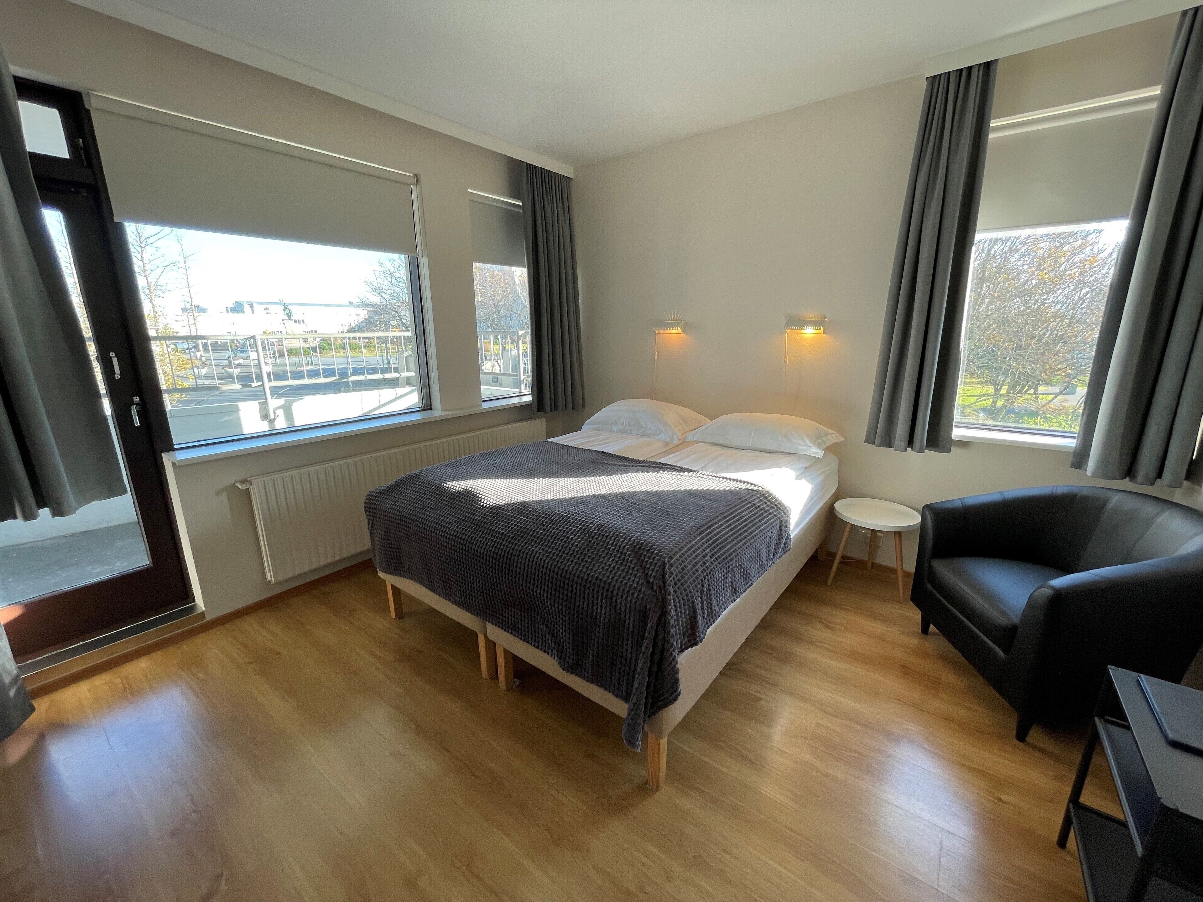 A modern hotel room with a large bed, two bedside tables, a black leather chair, and a view of the city through large windows.