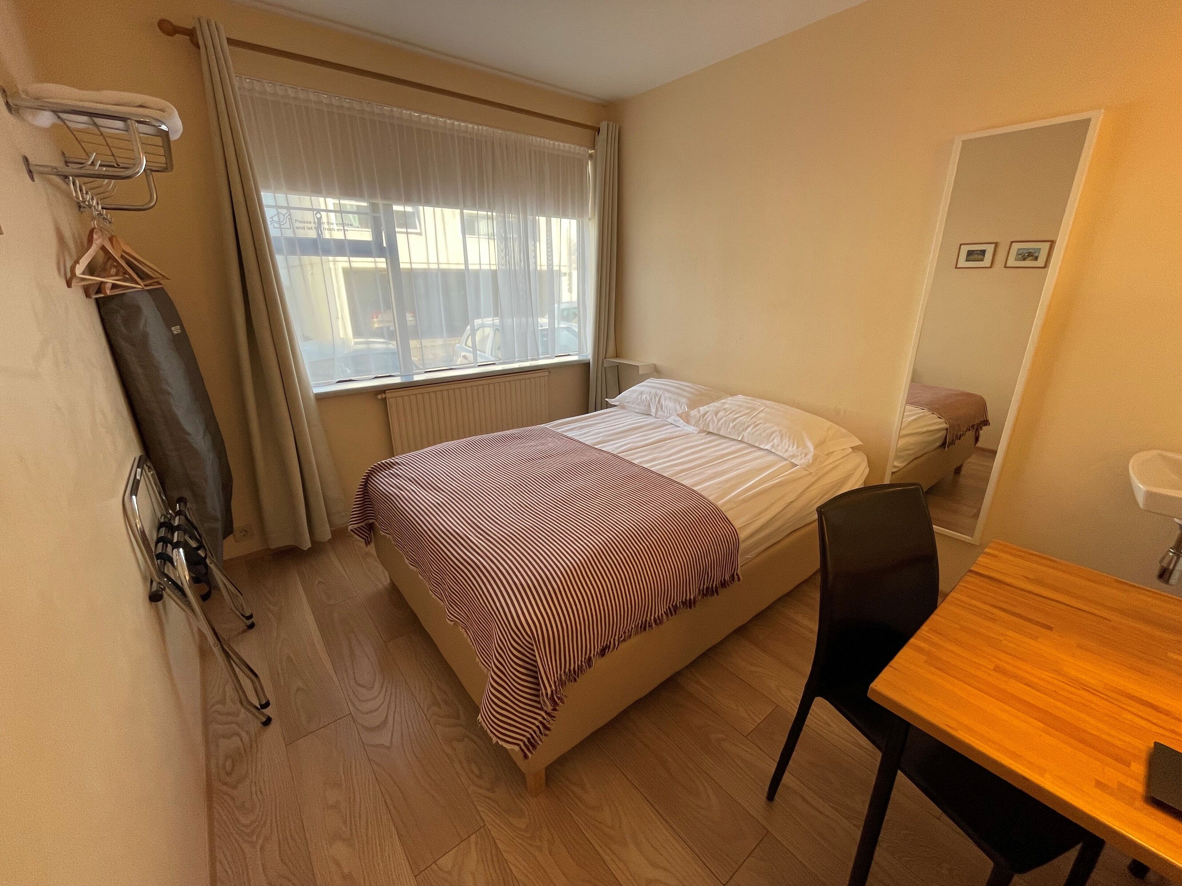 A hotel room with a bed, a wooden desk, a chair, a radiator, a window with curtains, a towel rack, and a small mirror.
