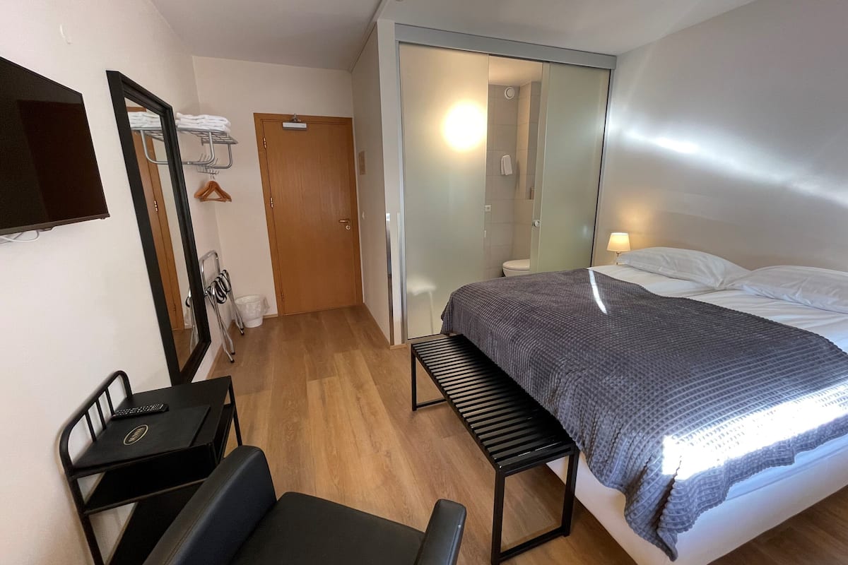 Quarto de hotel com uma cama, uma escrivaninha com cadeira, um banco, um espelho e uma porta para um banheiro.