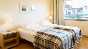 A hotel room with two beds, a bedside table with a lamp, a small shelf, and a window with a view of buildings. - Guesthouse Sunna (Reykjavik)