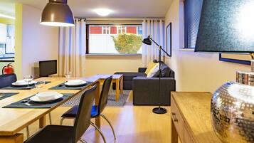 Moderni hotellihuone, jossa on ruokailutila, sohva ja suuri ikkuna, jossa on sälekaihtimet.