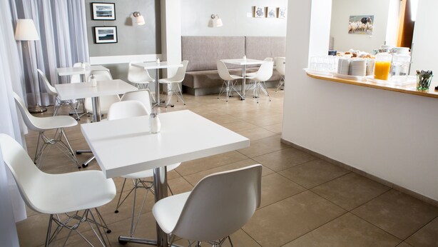 A modern dining area with white tables and chairs, a counter with breakfast items, and a grey cushioned seating area. - Guesthouse Sunna (Reykjavik)