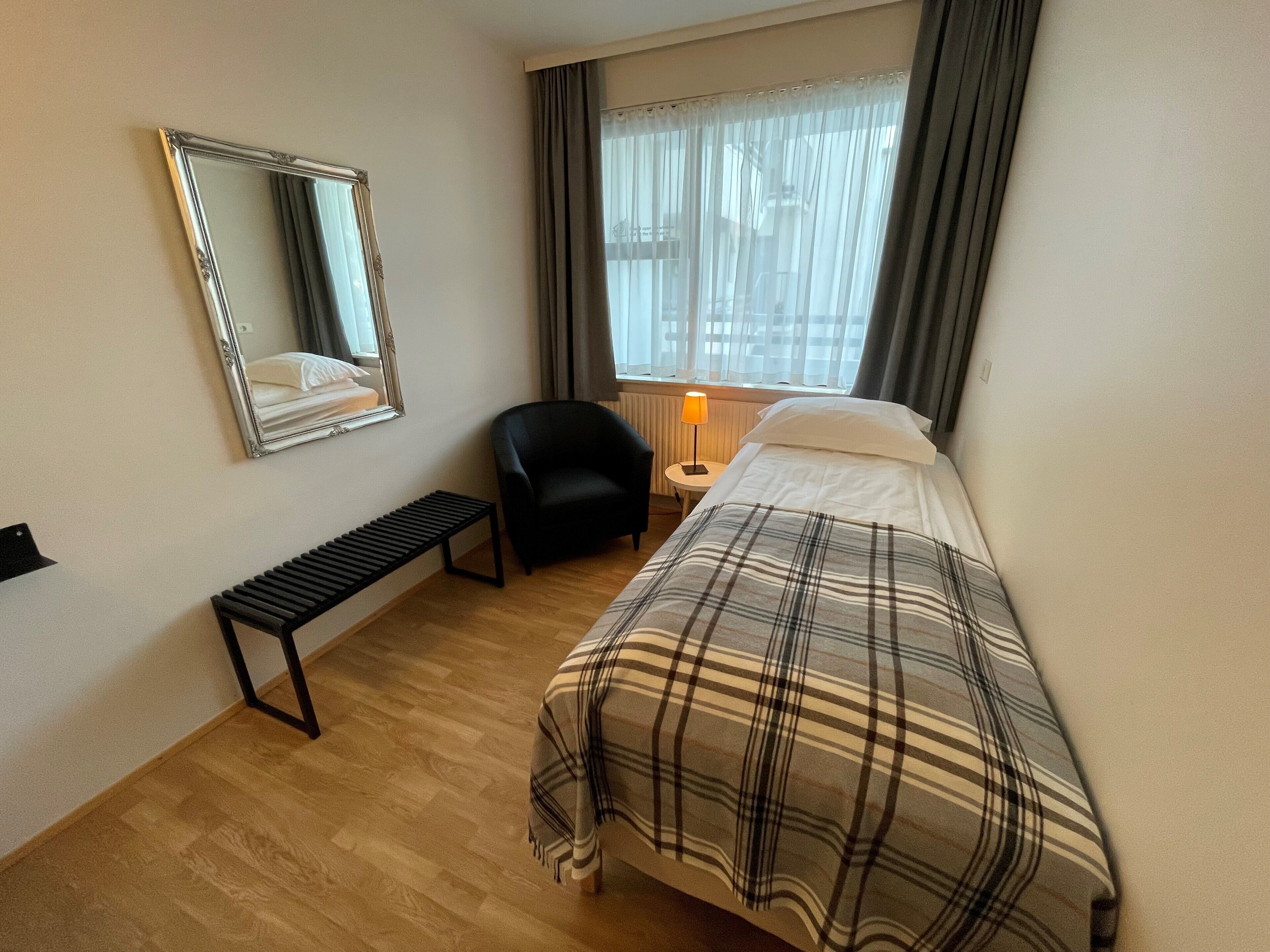 Een hotelkamer met een bed, een bankje, een stoel, een nachtkastje met een lamp, een raam met gordijnen en een spiegel aan de muur.