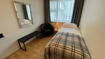 A hotel room with a bed, a bench, a chair, a bedside table with a lamp, a window with curtains, and a mirror on the wall.