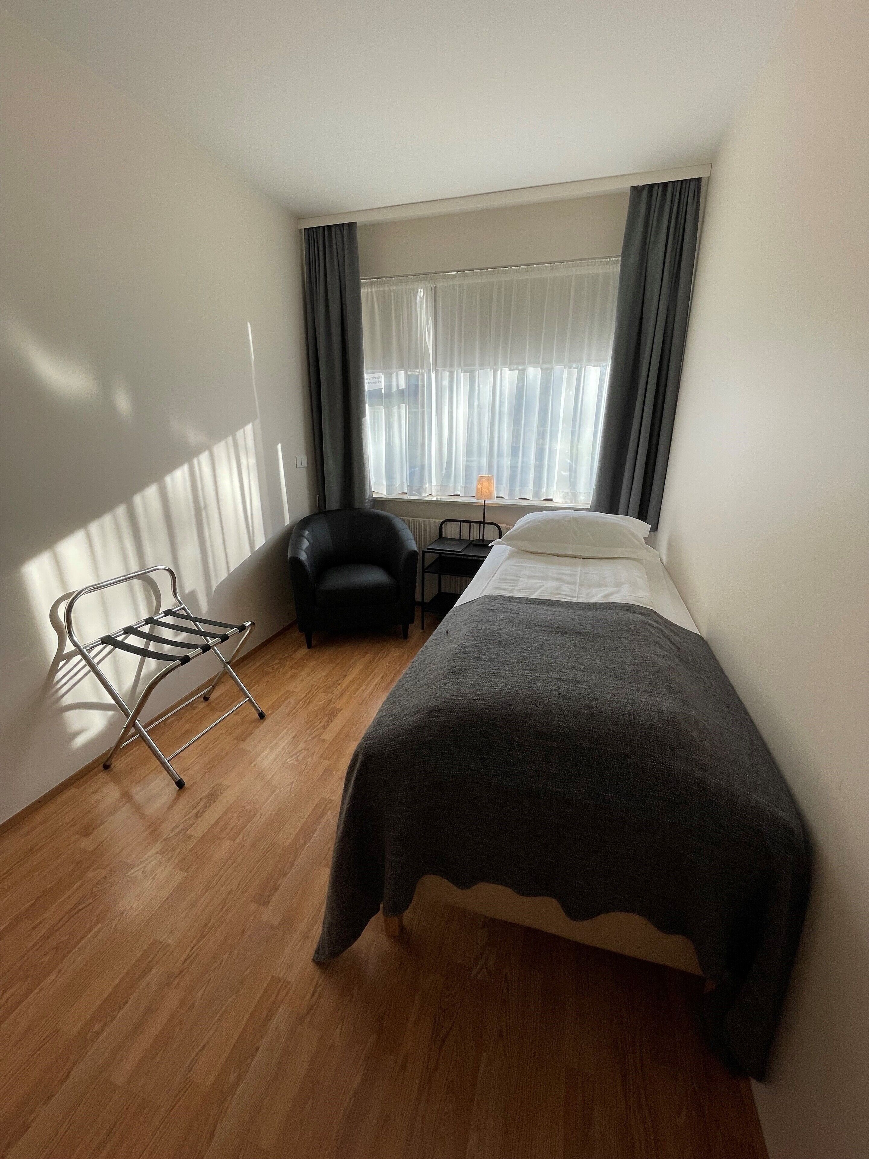 Un dormitorio pequeño y ordenado con cama, silla, escritorio y ventana con cortinas.