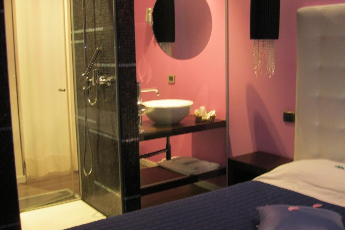 Een hotelkamer met een douchecabine van glas, een witte wastafel, een nachtkastje met een spiegel en een bed met een blauwe deken en roze bloemen.