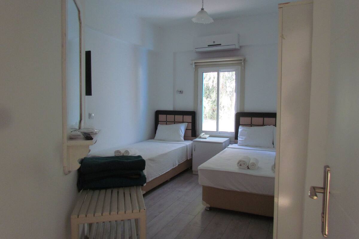 A hotel room with two single beds, a small table, a bench, a window, and an air conditioning unit.