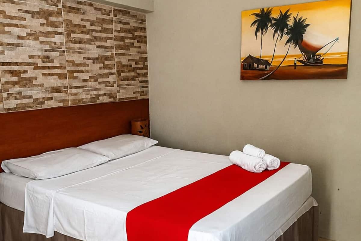 A hotel room with a bed, a red throw, and a painting of a sunset.