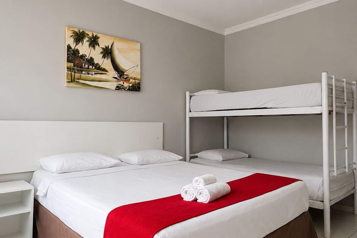 A hotel room with a bunk bed, a single bed, a red throw blanket, and a painting of a beach scene.