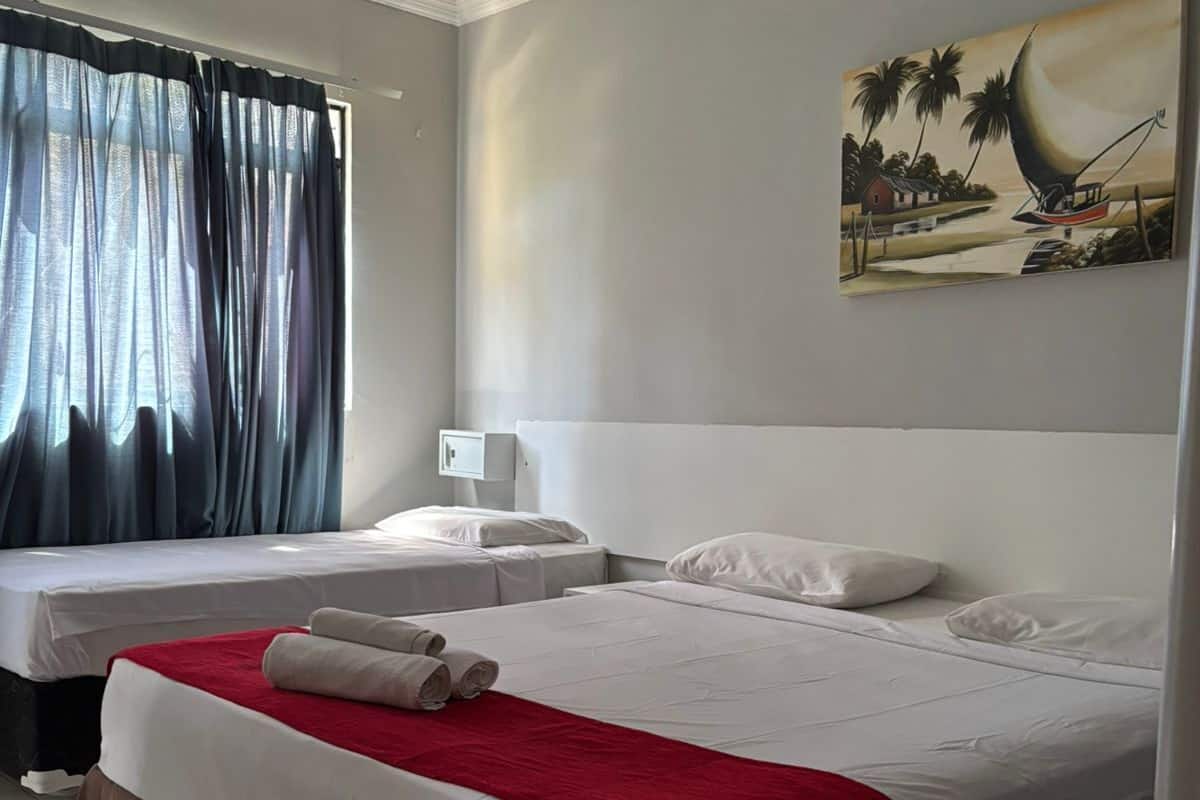 A hotel room with two beds, a red and white bedspread, a painting on the wall, and a window with curtains.