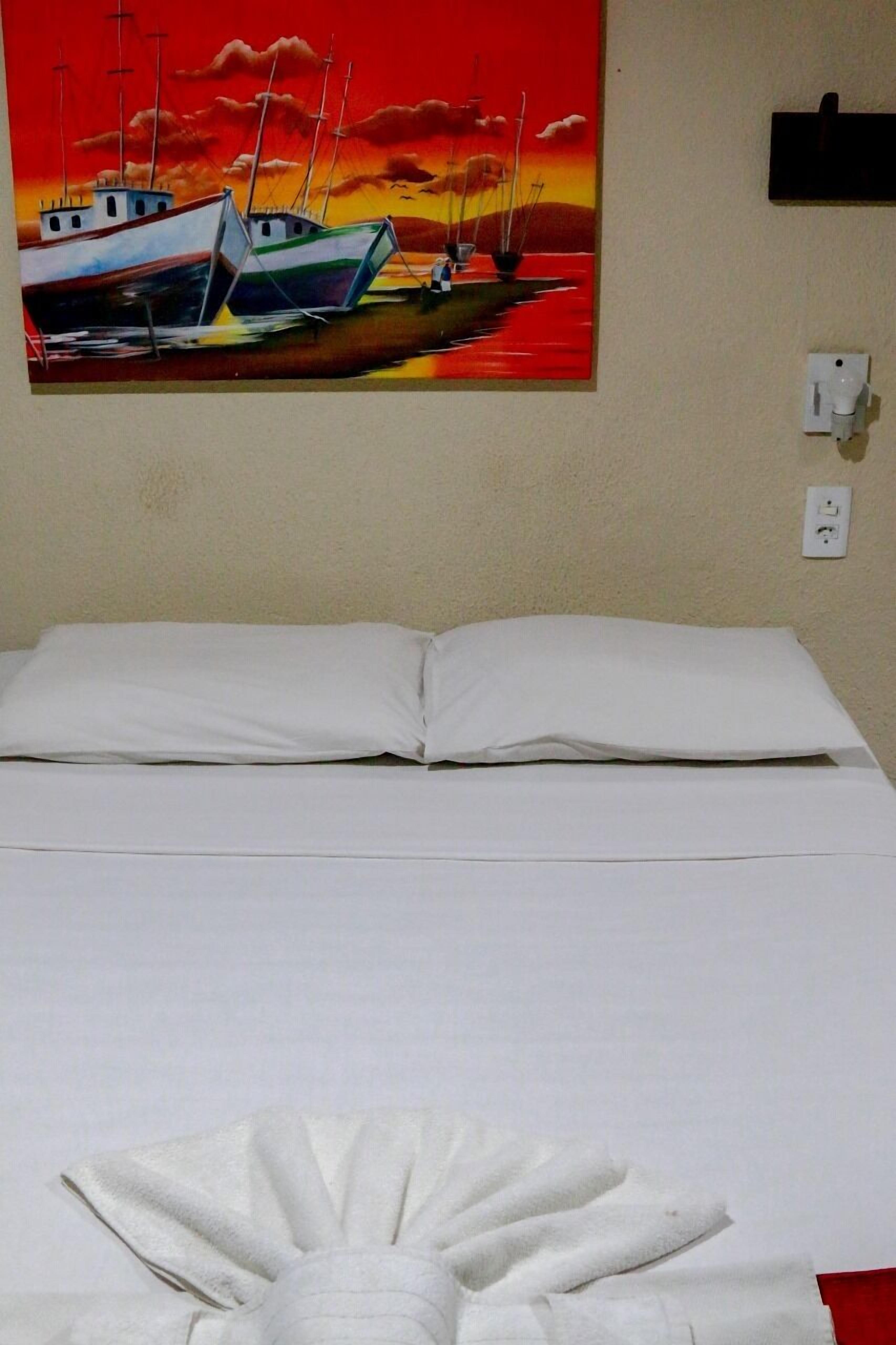 A bed with white linens, a painting of boats on a sunset, and a wall socket.