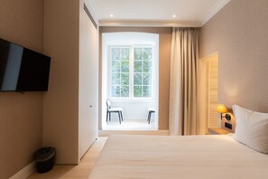 Ein Hotelzimmer mit Bett, Fernseher, einem Fenster mit Vorhängen, einem Sessel und einem Nachttisch mit Lampe.