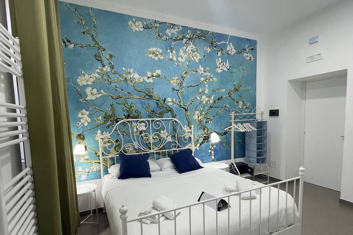 A neatly arranged bedroom with a white metal bed frame, a blue floral wall mural, and a small bedside table with a lamp.
