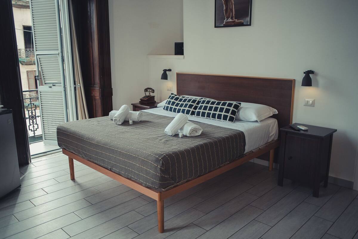 A neatly made bed with a wooden frame and a grey striped bedspread. There are two white pillows and two rolled towels on the bed. A small bedside table with a lamp and a remote control is to the right of the bed.