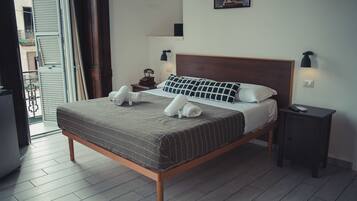 Una cama bien hecha con estructura de madera y una colcha a rayas grises. Hay dos almohadas blancas y dos toallas enrolladas sobre la cama. A la derecha de la cama, una mesita de noche con una lámpara y un control remoto.