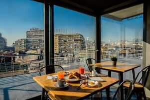 Ontbijttafel met croissants, eieren en sap, met uitzicht op een stadsgezicht met gebouwen en een zeezicht.