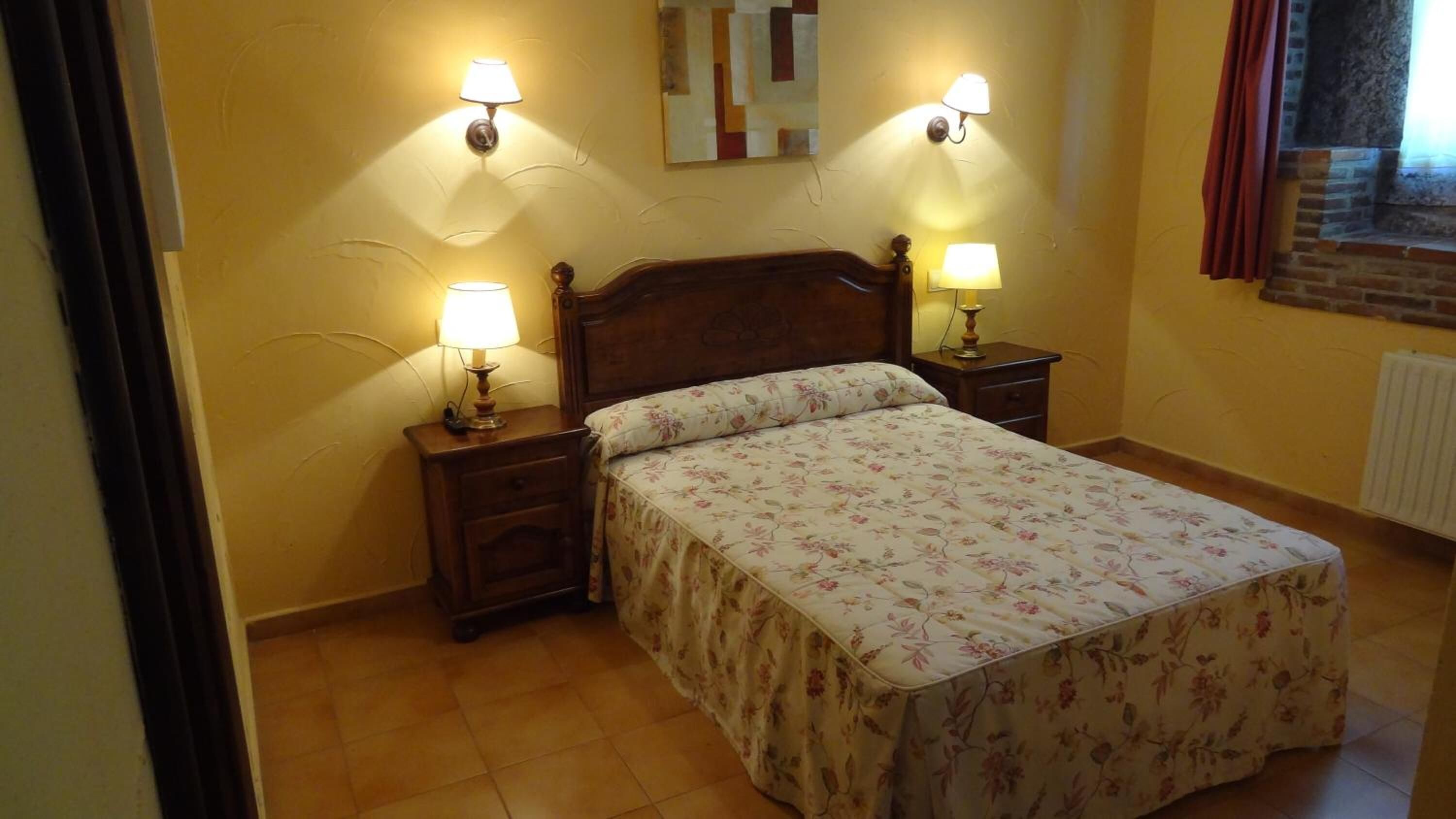 Un dormitorio con cama de madera, ropa de cama floral, dos mesitas de noche con lámparas y un espejo fijado a la pared.