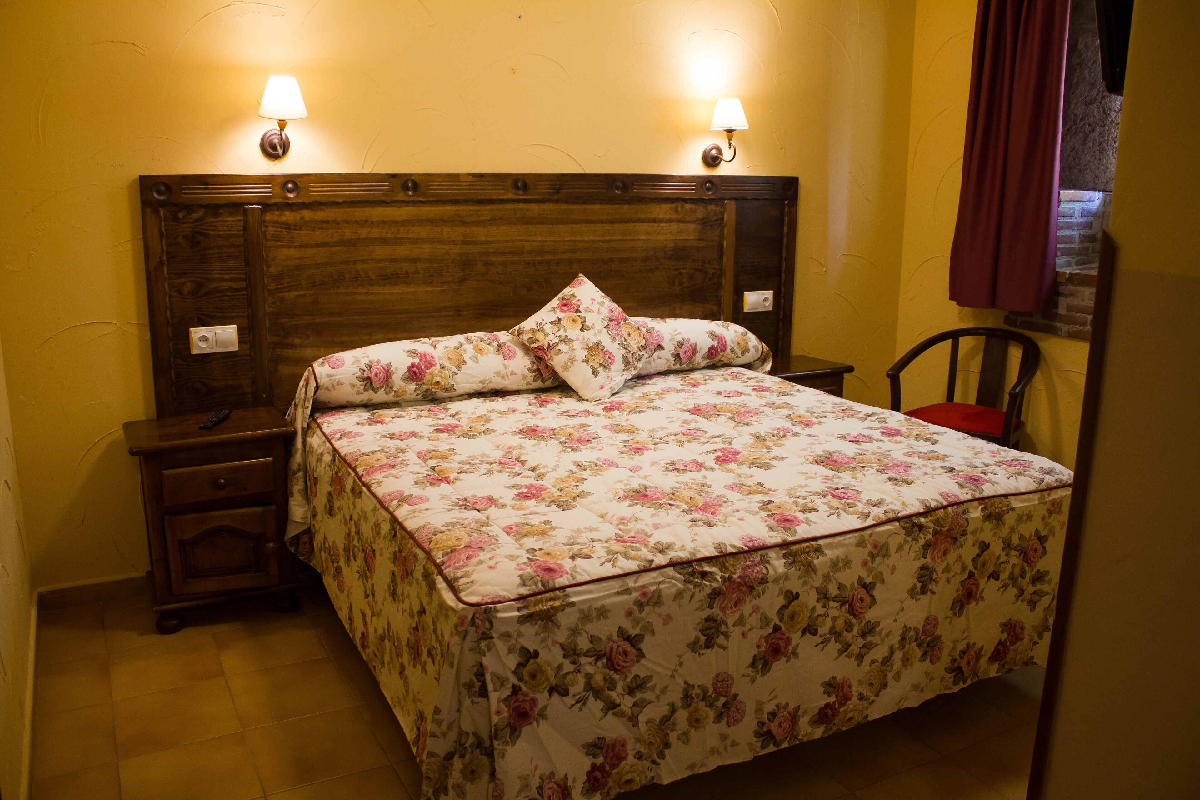 Una cama bien tendida con ropa de cama floral, dos lámparas de noche, una mesita de noche y una silla.