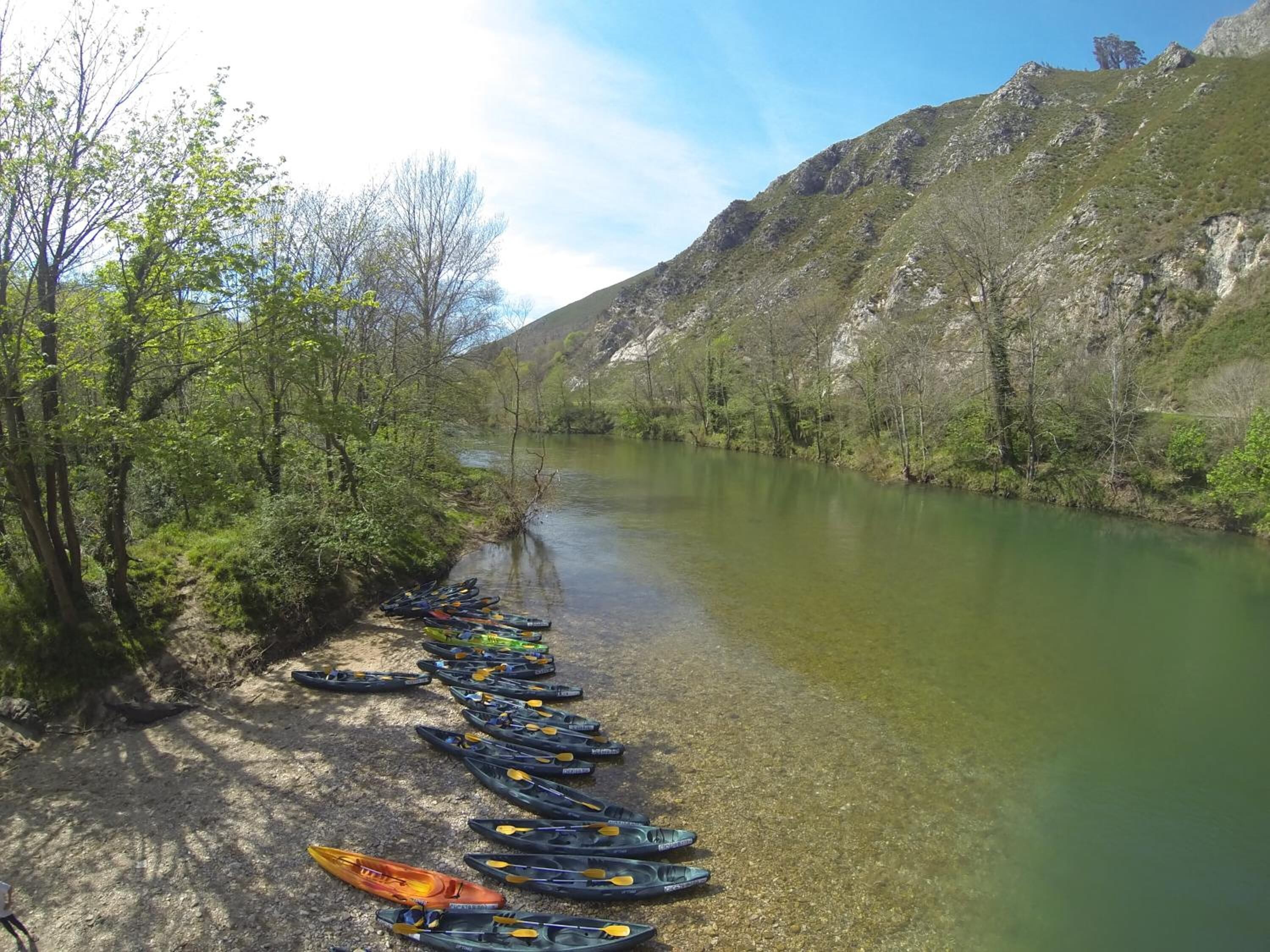 Un río con kayaks alineados en la orilla, rodeado de árboles y colinas.