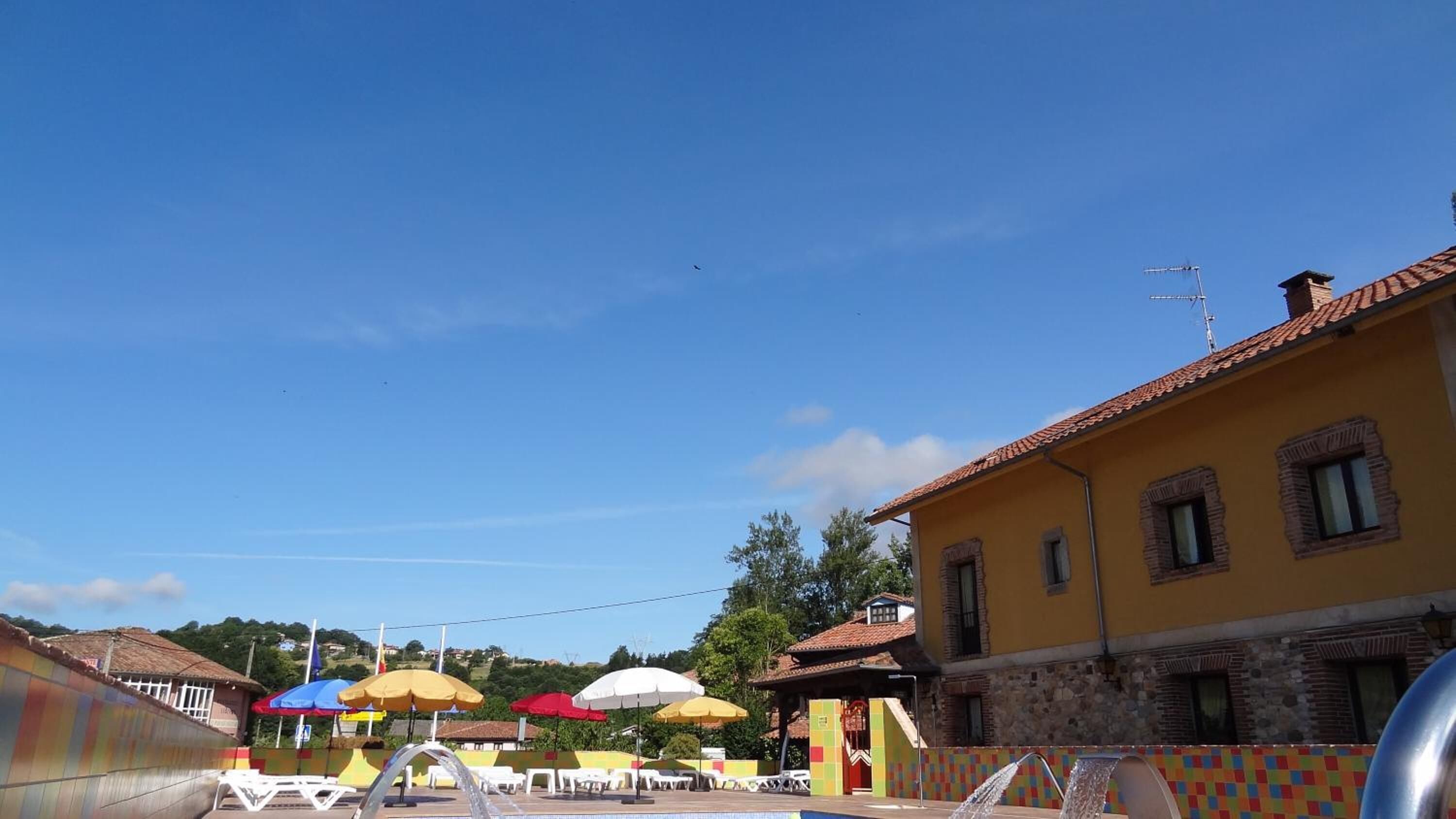 A pool area with lounge chairs and umbrellas, a yellow building with stone walls, and a clear blue sky.