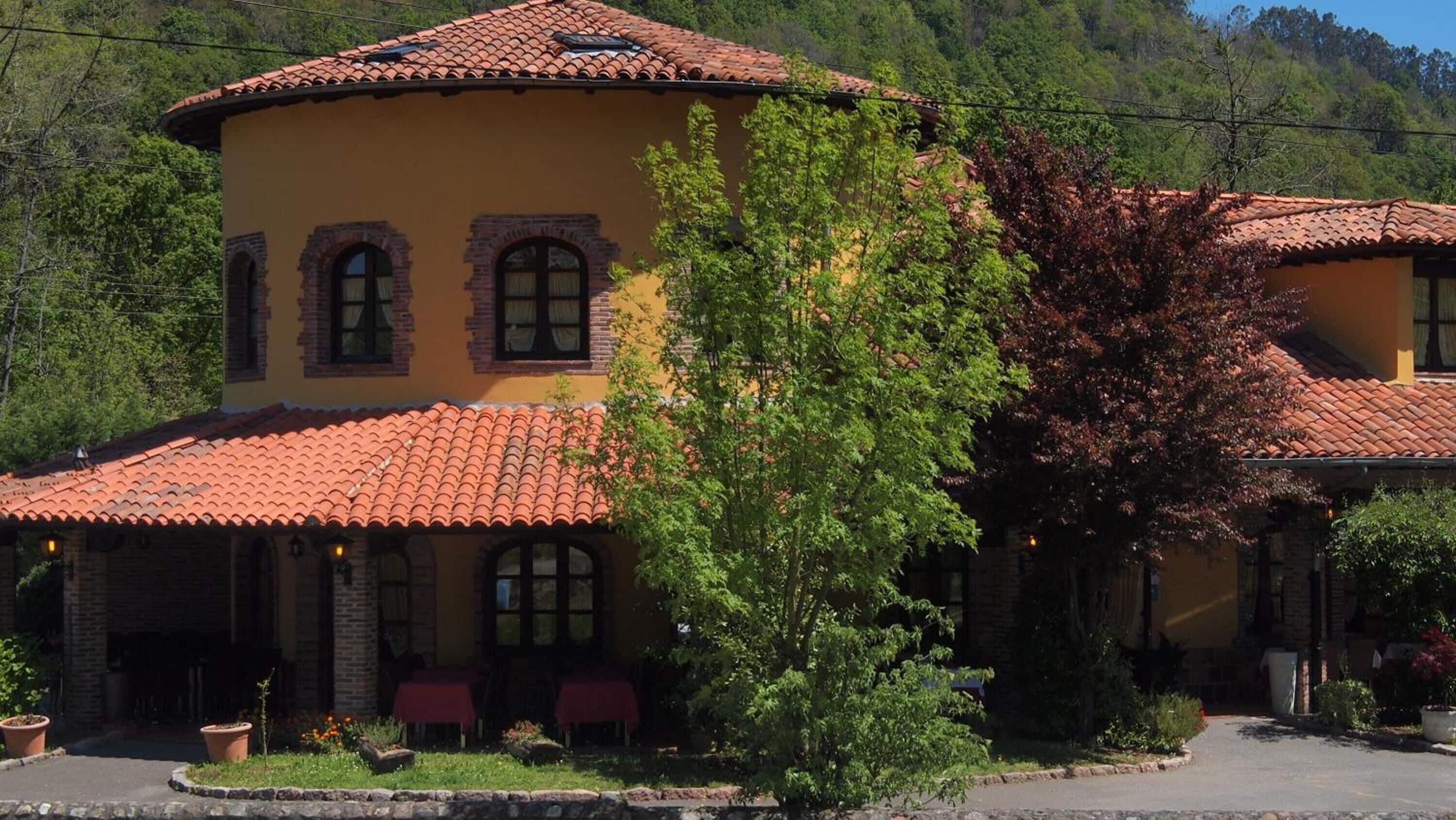 A two-story building with a red-tiled roof, arched windows, and a garden area with potted plants.