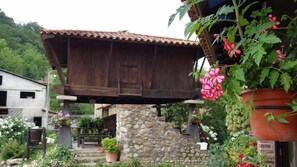 A rustic wooden structure with a tiled roof, supported by stone pillars, surrounded by lush greenery and flowering plants.