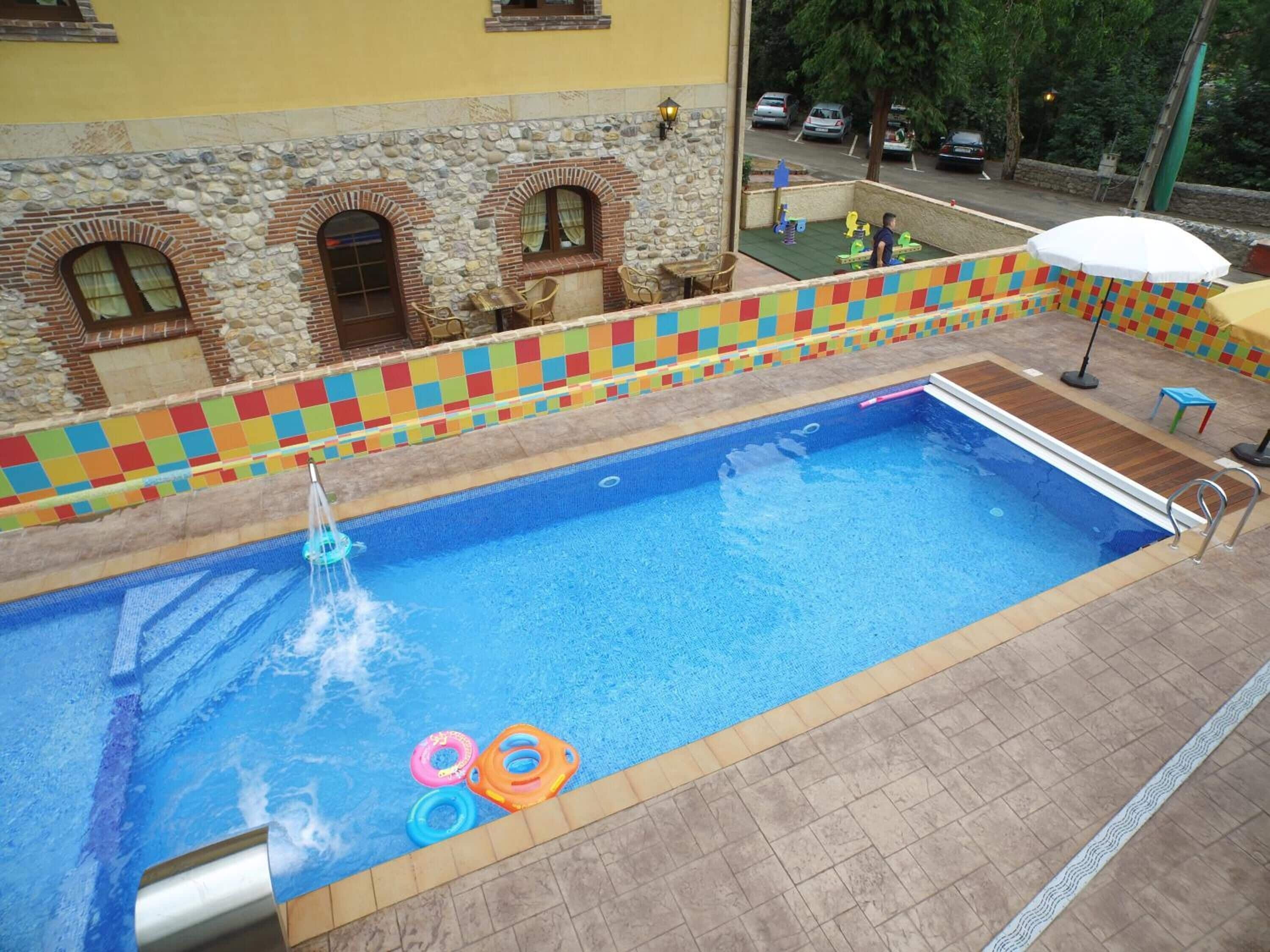 A swimming pool with a stone wall and colorful tiles, surrounded by a tiled deck and a few chairs.