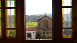 Vista a través de un ventanal con vidrieras, mostrando un edificio de piedra con tejado rojo y una cruz en la parte superior, sobre un paisaje verde.