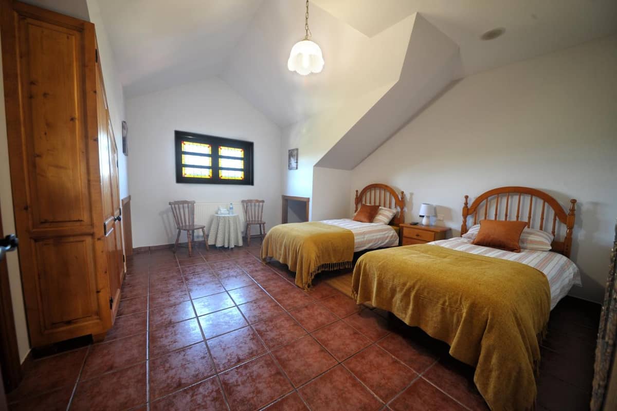 A bedroom with two beds, a wooden wardrobe, a small table, and a window with stained glass.