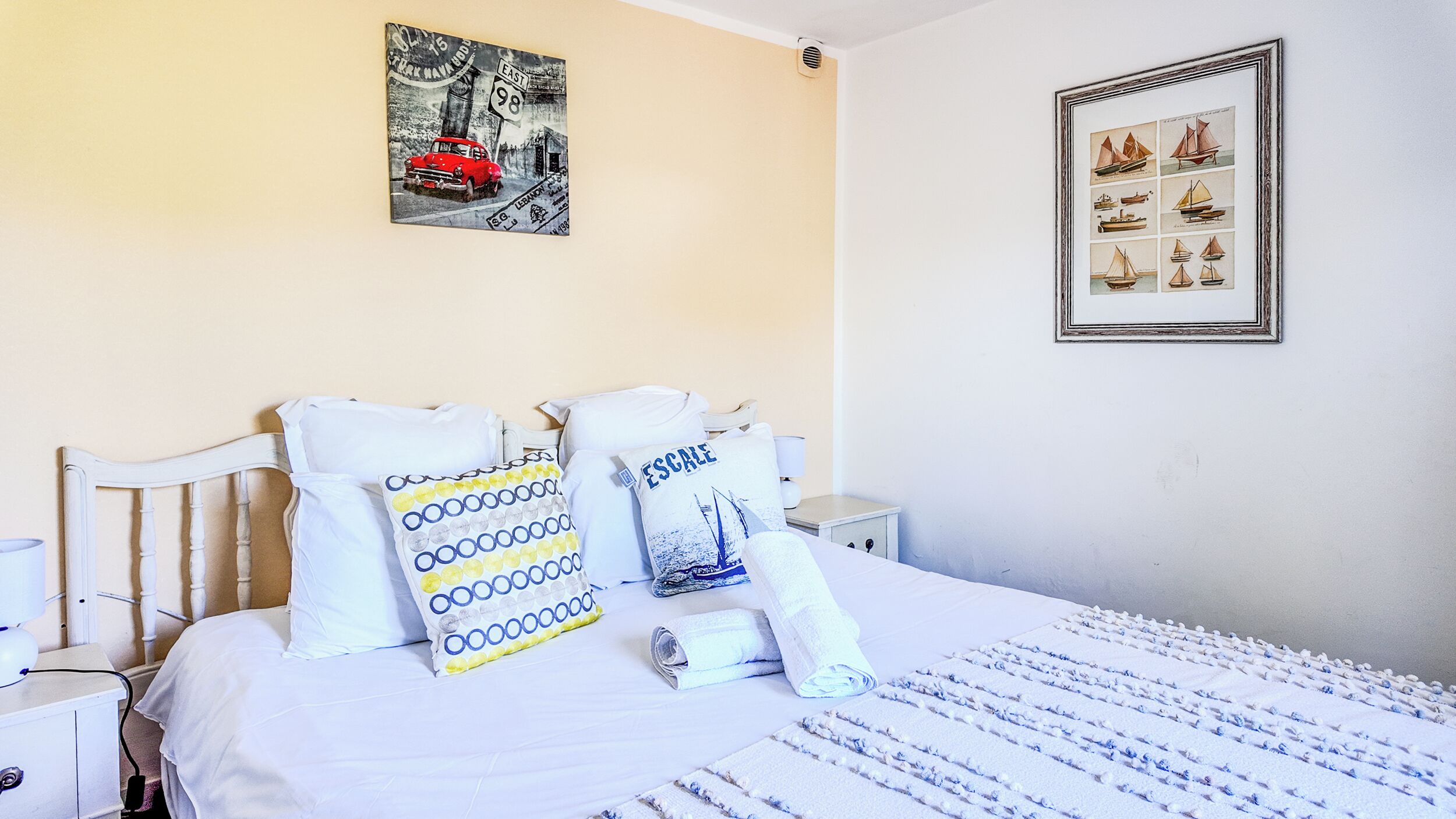 Una cama bien hecha con ropa blanca y almohadas estampadas, un cuadro enmarcado de veleros en la pared e ilustración de un coche antiguo sobre la cama.