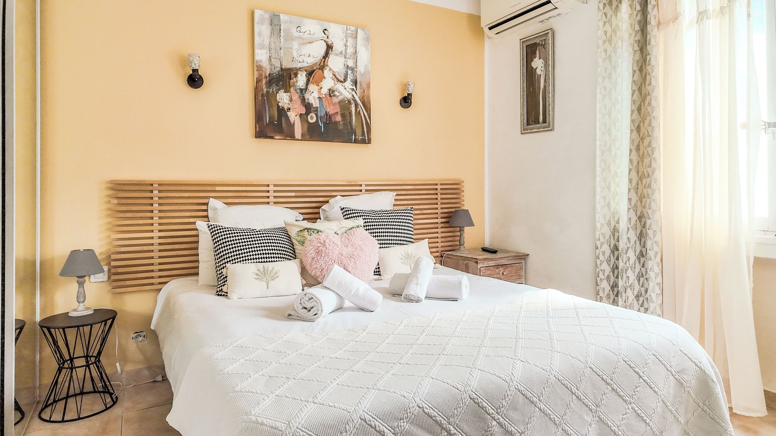 A neatly made bed with a patterned quilt, a wooden headboard, and various pillows. There are two bedside tables with lamps, and a painting above the bed.
