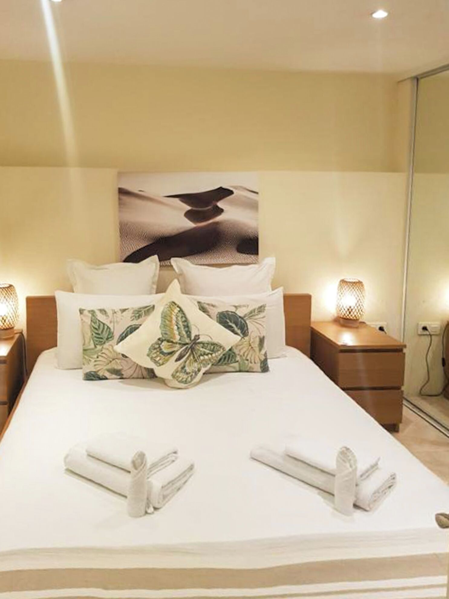 A neatly made bed with white linens, two pillows, and a tropical leaf-patterned pillow. There are two bedside lamps and a painting of a bird on the wall.
