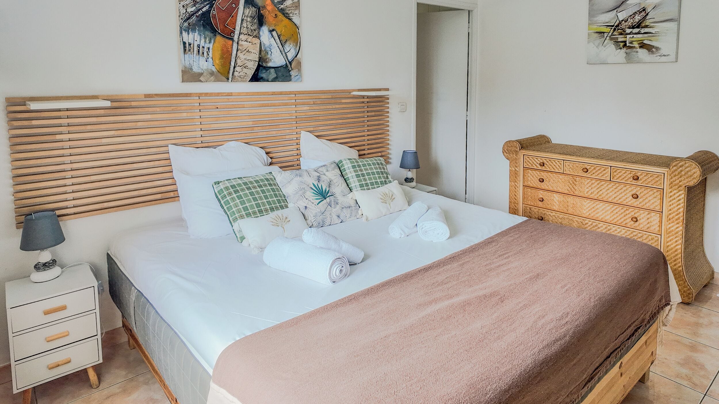 Un lit bien fait, avec une couverture rose, une tête de lit en bois et plusieurs oreillers. On y trouve une petite table de chevet avec une lampe et un tiroir.