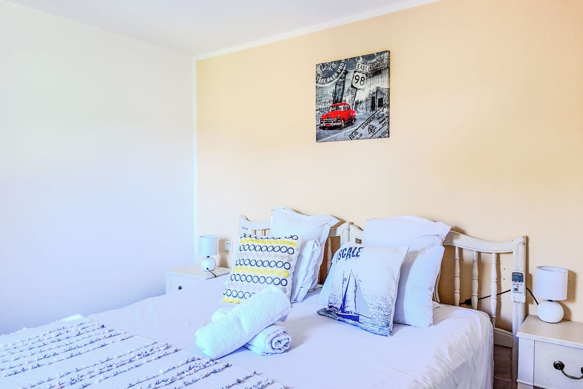 A neatly made bed with white linens and a patterned pillow. There is a small bedside table with a lamp and a wall-mounted artwork depicting a vintage car and a street sign.