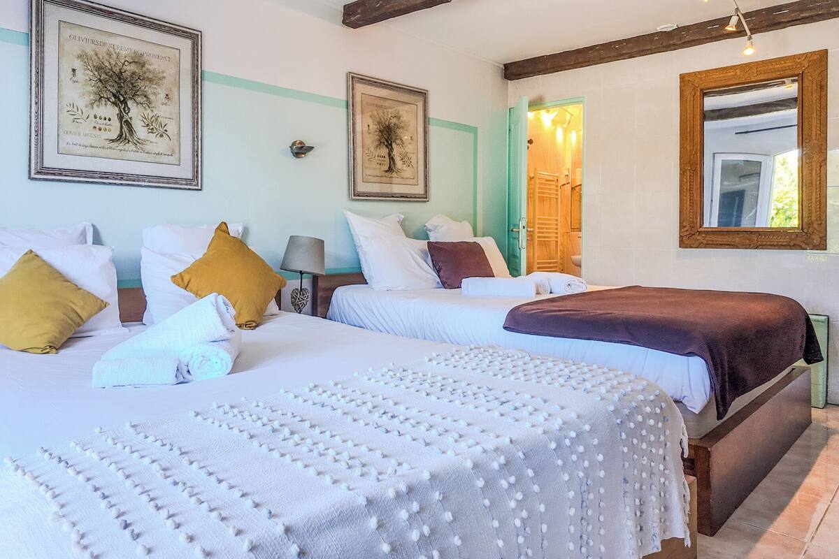 A hotel room with two beds, each with white bedding and decorative pillows. There are framed pictures on the wall, a wooden headboard, and a mirror.