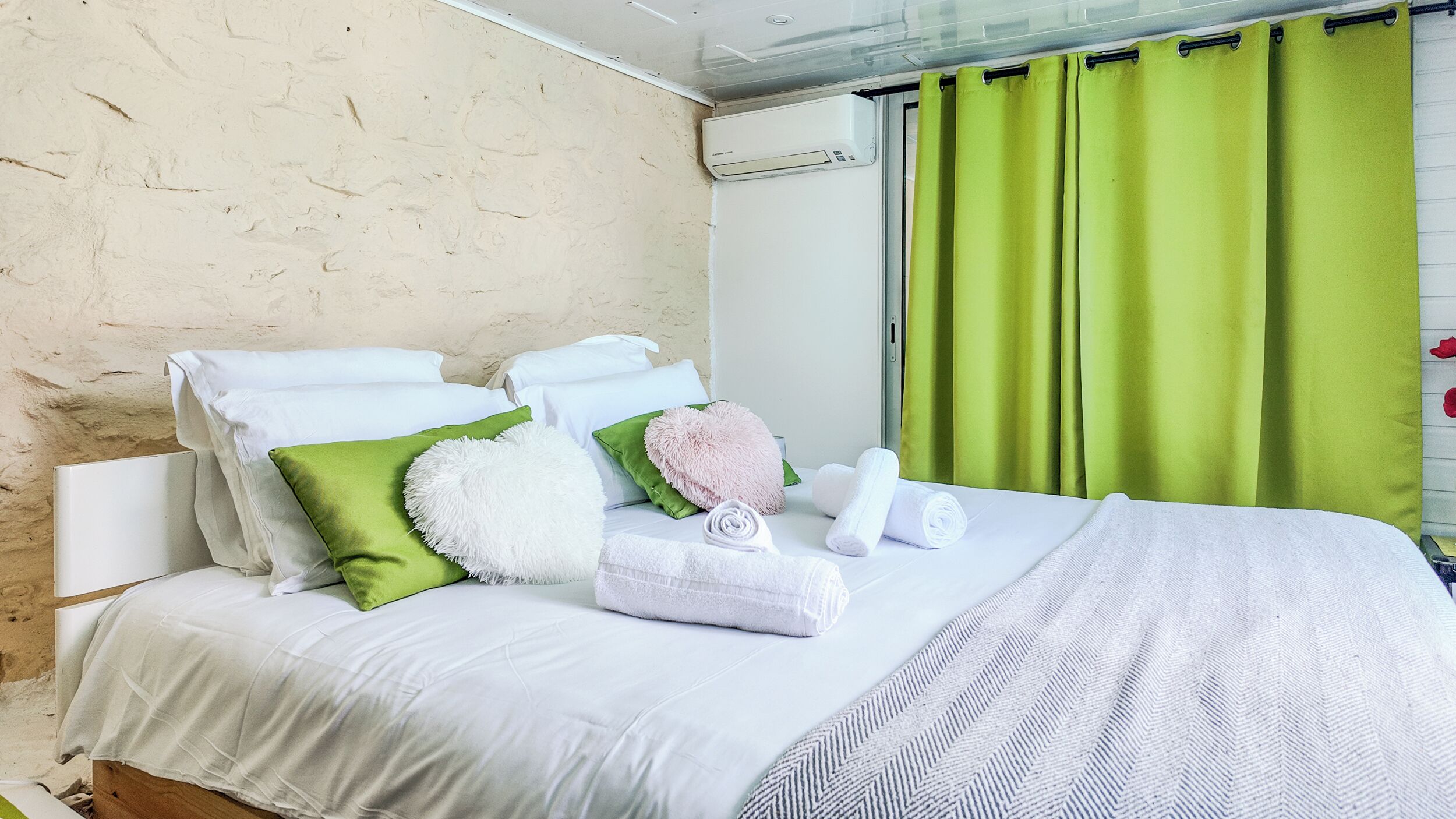 A neatly made bed with white linens, green and pink pillows, and a rolled towel.