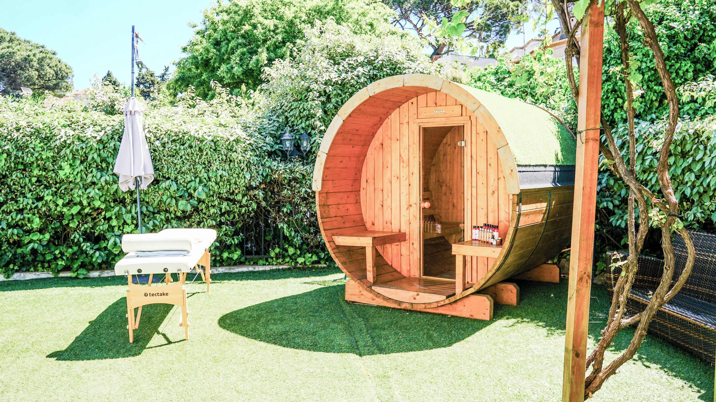 A wooden sauna with a bench and a table, surrounded by a hedge and a grassy area.