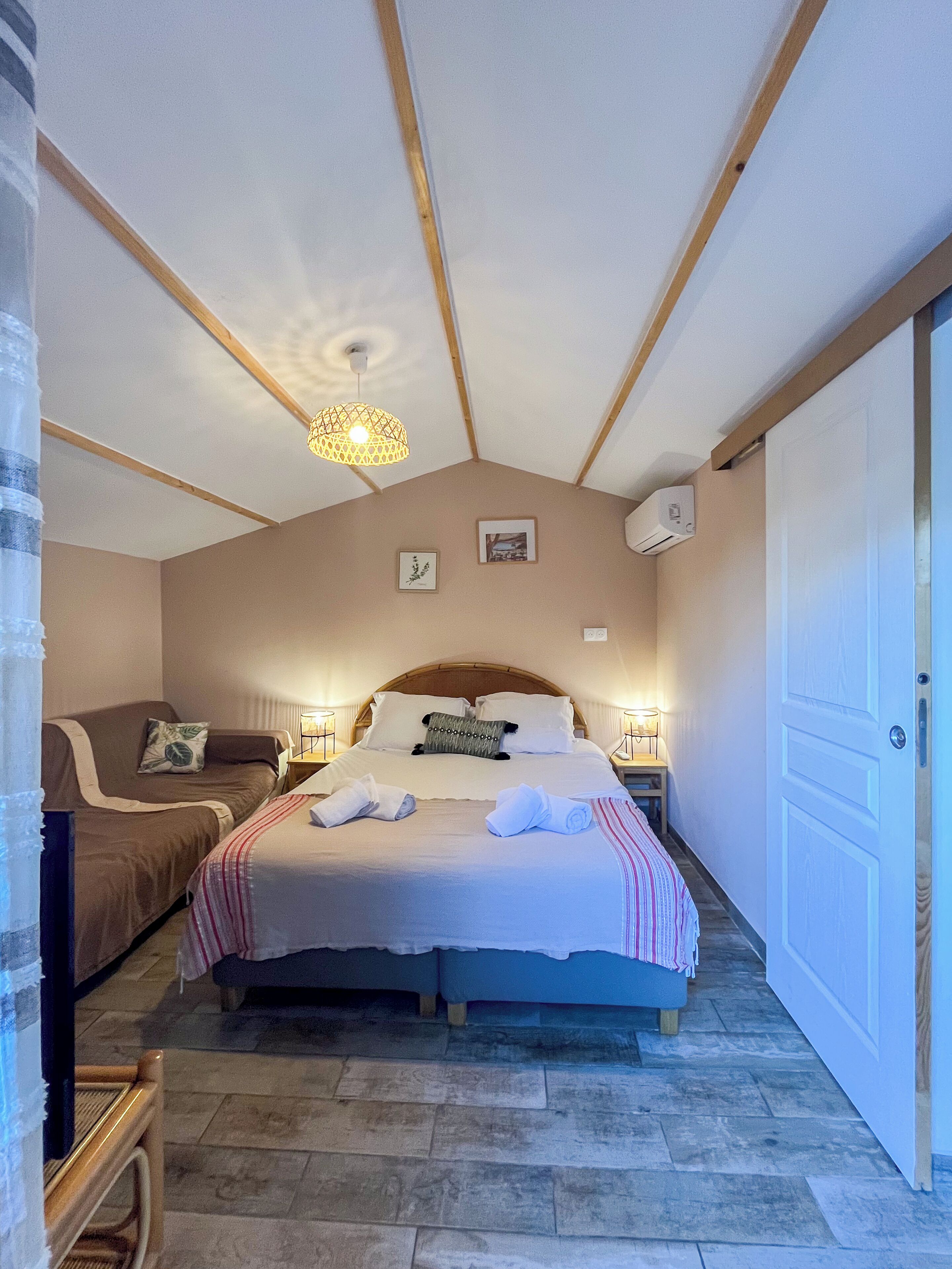 A bedroom with a large bed, a smaller sofa, a wooden ceiling, and a door.