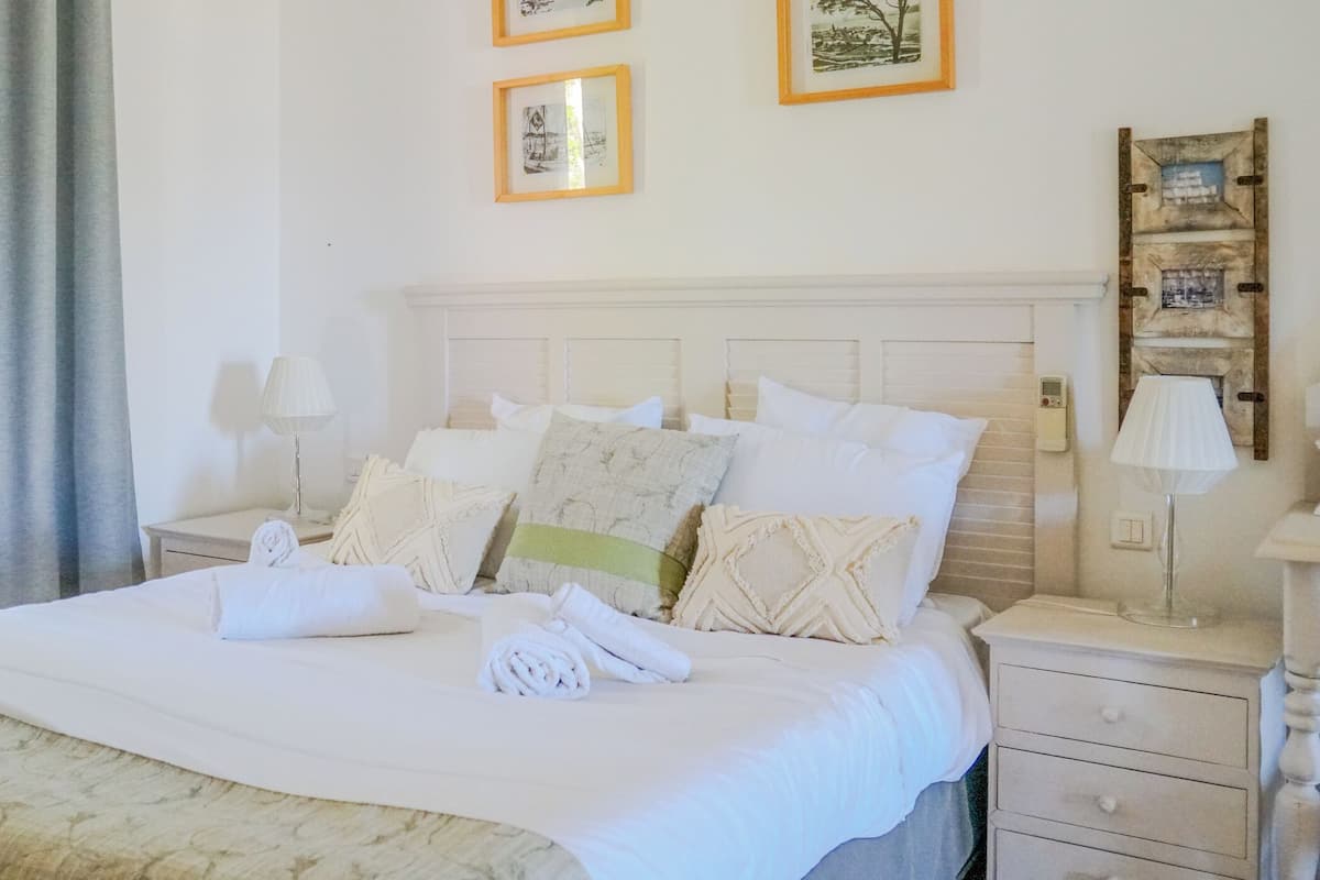 A neatly made bed with multiple pillows and a blanket, a bedside lamp, a nightstand, and a wall-mounted shelf with framed pictures.
