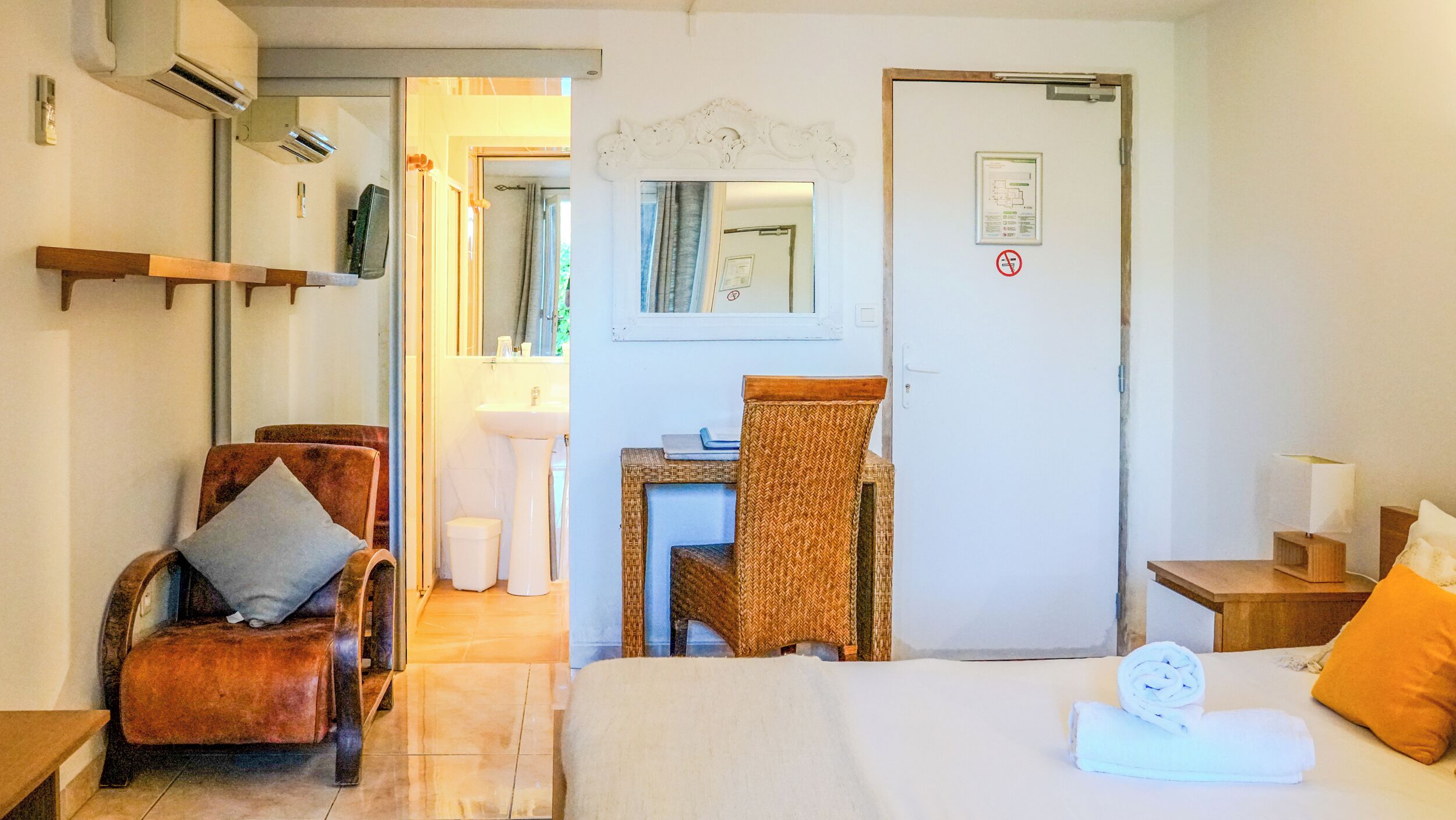 Hotellihuone, jossa on sänky, tuoli, pieni pöytä, peili ja kylpyhuone, joka näkyy avoimesta ovesta.