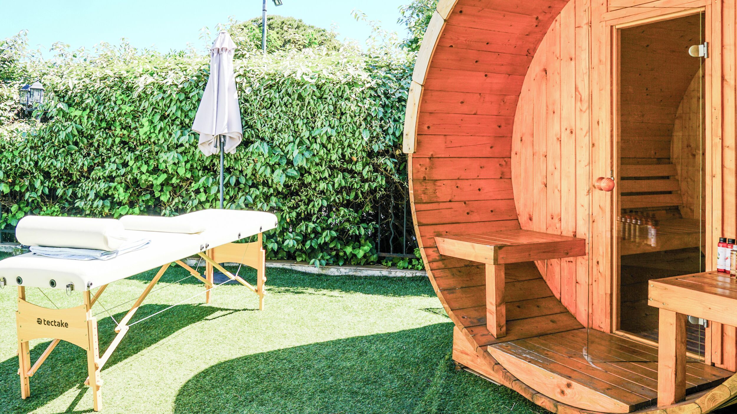 Una sauna de madera con un banco y una mesa, rodeada por un seto cuidado y un césped.