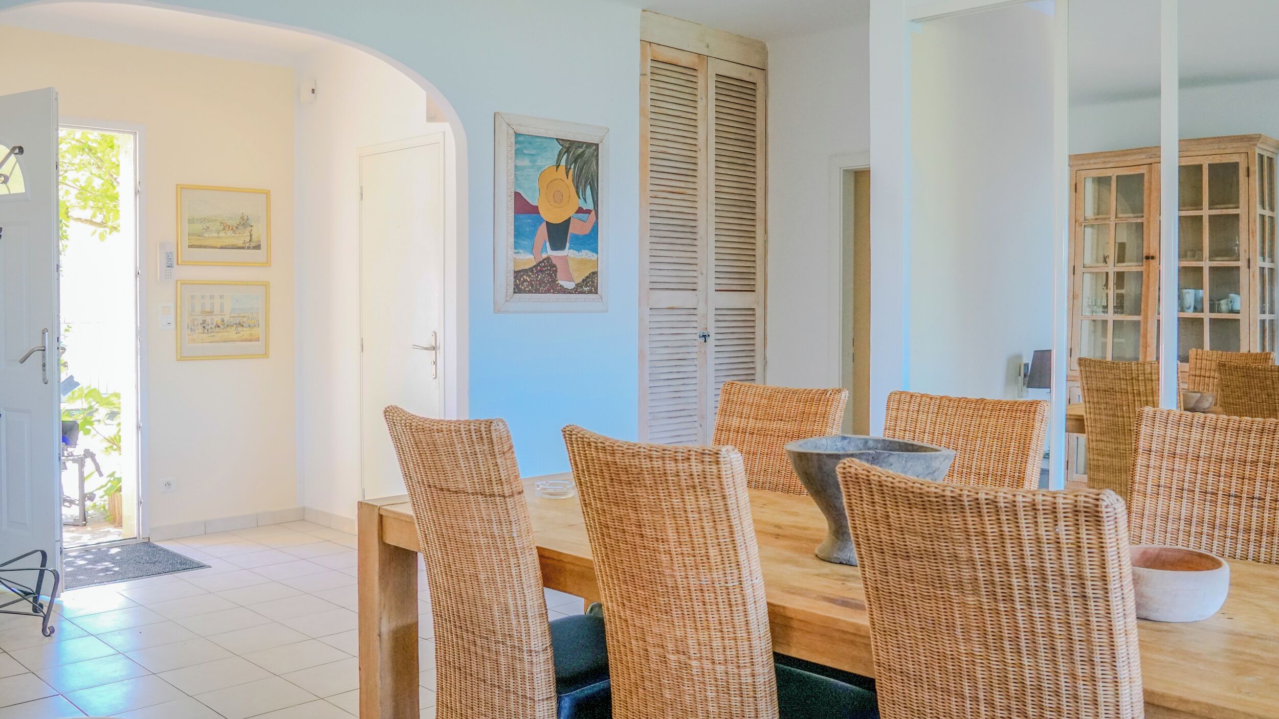 A dining area with wicker chairs and a wooden table, a door leading outside, and a wall with framed artwork.