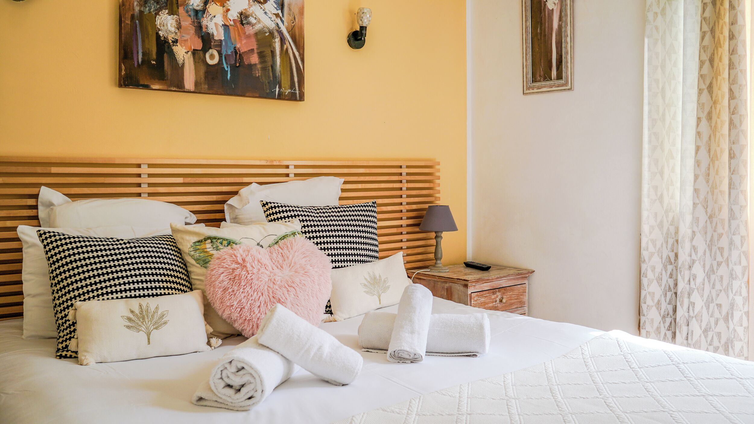 A neatly made bed with white linens and multiple pillows, a wooden headboard, a bedside table with a lamp, and a painting on the wall.