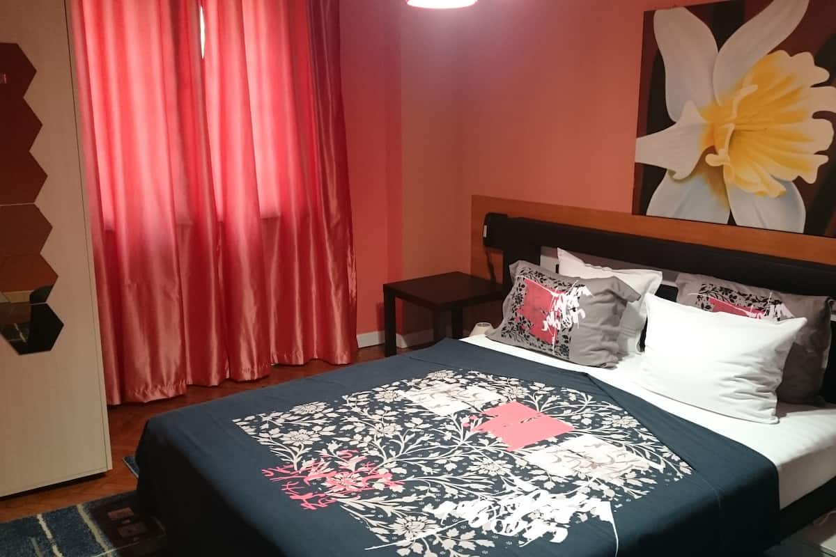 A bedroom with a bed, pillows, a nightstand, a lamp, and a large floral artwork on the wall.