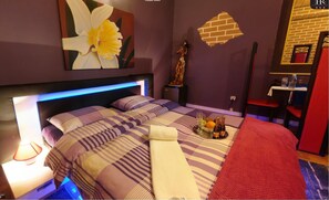 A hotel room with a bed, a nightstand, a tray with fruits and drinks, and a red chair.