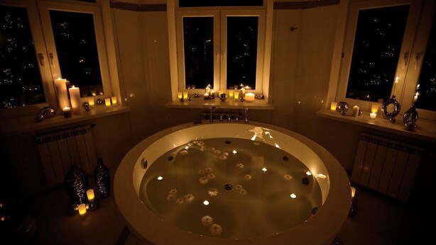 A large, oval-shaped hot tub with candles floating on the water, surrounded by windows with candles on the windowsills. - Dom Aktera (Odesa)